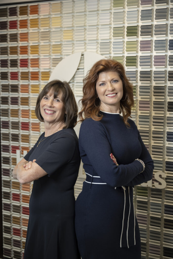 Two women stand back-to-back, smiling, in front of a wall covered with rows of colored samples.